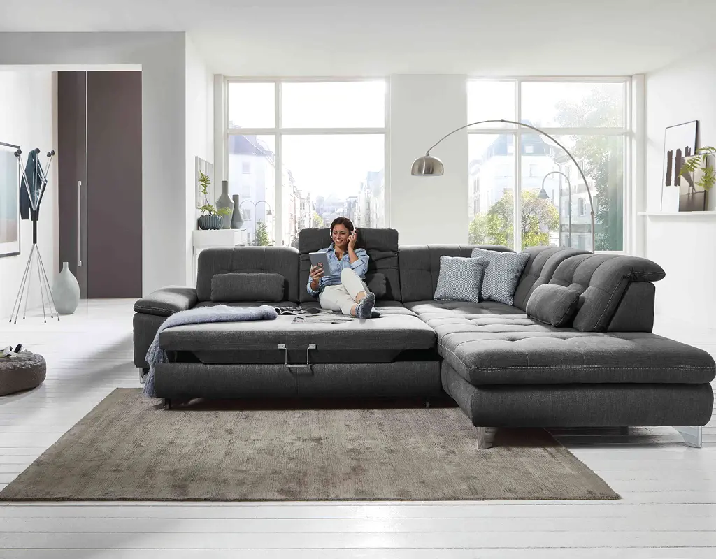 Großes graues Ecksofa mit Relaxfunktion in hellem Wohnzimmer. Frau sitzt entspannt auf dem modernen Polstermöbel. Komfortable Wohnlandschaft für gemütliche Stunden im Zuhause.